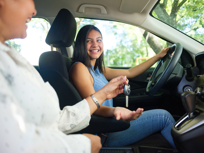 A side view photo of a teenage girl in a car as her mother sits next to her and hands her the keys to drive.